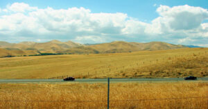 a golden valley of grass with rolling hills, a blue sky and white clouds in the background with a road and two cars in the foreground