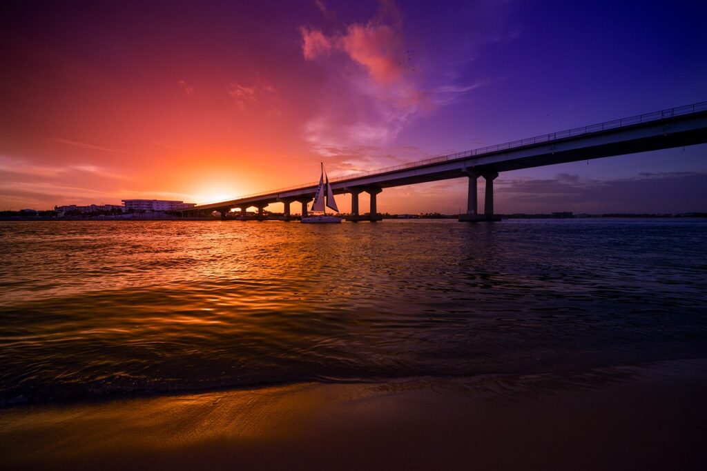 orange and purple sunset with the silhouette of a bridge over the ocean with a sailboat