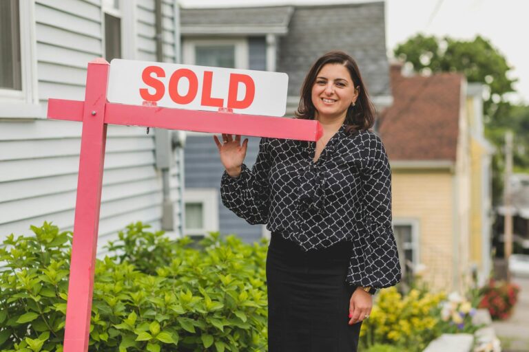 Smiling real estate agent with dark hair standing outside of a white home with a sold sign on an orange stand